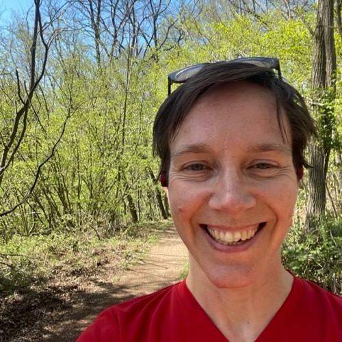Eine lächelnde Frau mit kurzen, braunen Haaren und einer Brille auf dem Kopf steht auf einem Weg im Wald.