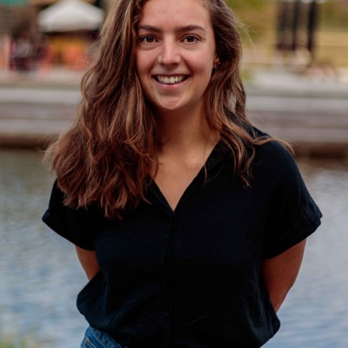 Portrait photo. Sanne is wearing a black blouse and jeans. In the background you see slightly some water and a green environment like a parc. 