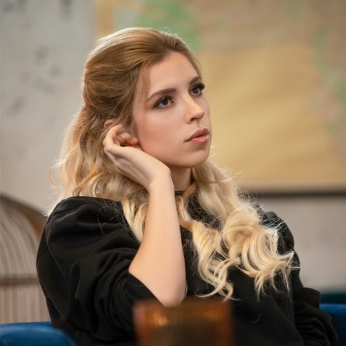 Auf dem Foto sieht man Shurjoka. Sie wurde bei einem Dreh auf dem Set fotografiert und schaut verträumt an dem Fotografierenden vorbei. Sie hat blonde gelockte Haare, mit der rechten Hand nestelt sie an ihrem Ohr und trägt einen schwarzen Hoodie. Das Bild hat eine rustikale Vintage Atmosphäre. 