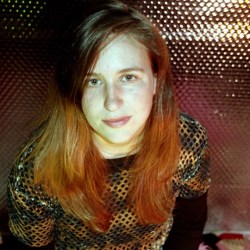photo of a white person with long hair looking directly at the camera, wearing a bleached mesh shirt over a black shirt, the background is a reflected metallic patterned surface